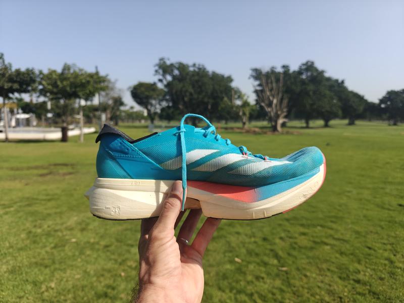 A hand holds up a pair of Adidas Adizero Adios Pro 3 running shoes in bright turquoise blue with white three-stripe logo, coral-orange midsole accents, and cream-colored sole, photographed outdoors in a park with green grass and trees under a clear blue sky