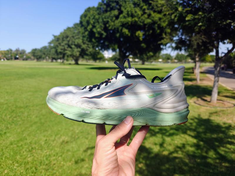 A hand holds up an Altra Torin 6 running shoe with light mint green upper, navy blue laces and mint green sole, photographed outdoors in a sunny park with green grass and large trees in the background