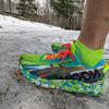 Close-up of my foot wearing a brightly colored ASICS Noosa Tri 13 running shoe in neon yellow-green with blue accents and red laces, standing on the ground with snow-covered forest terrain and birch trees visible in the background