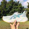 A hand holds up a white ASICS running shoe with blue side logos and an orange outsole, photographed outdoors in a sunny green park.