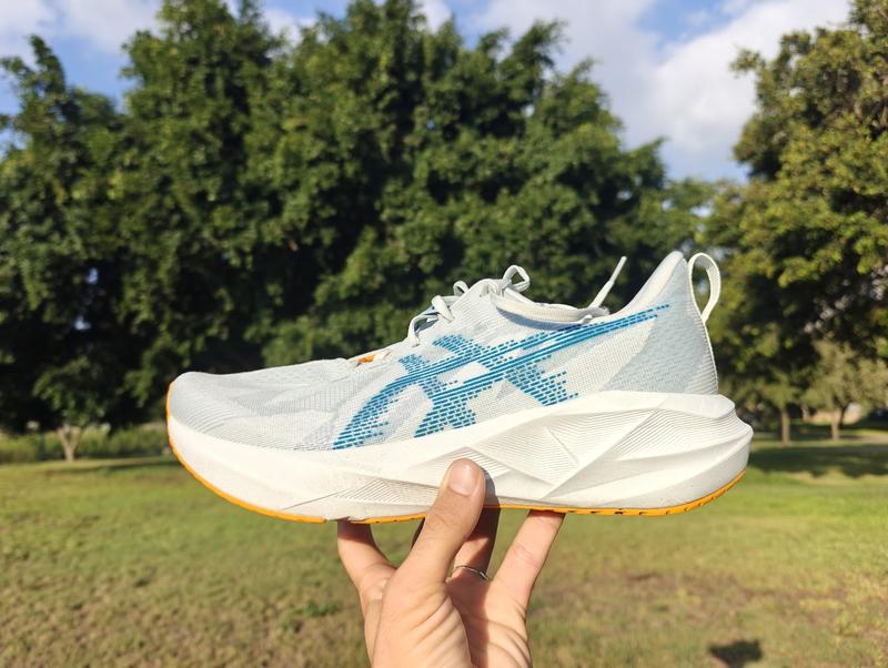 A hand holds up a white ASICS running shoe with blue side logos and an orange outsole, photographed outdoors in a sunny green park.
