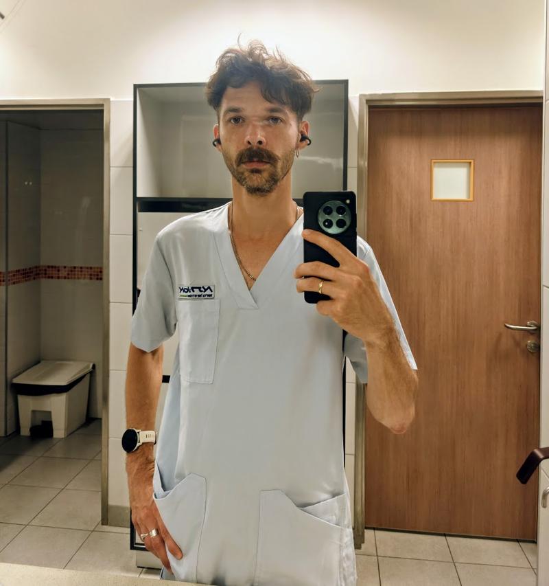 Mirror selfie of a hospital support worker wearing light blue scrubs in a staff restroom. He is holding a smartphone, wearing wireless earbuds and a smartwatch, with a neutral expression. Behind him are a wooden door, a wall mirror, and an open restroom stall with a trash bin. The photo is brightly lit and has a clean, clinical atmosphere.