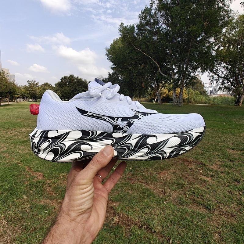 White Mizuno Wave Rider 30 with a black-and-white swirl-patterned midsole, held up on an open palm in a sunny park with tall eucalyptus trees.